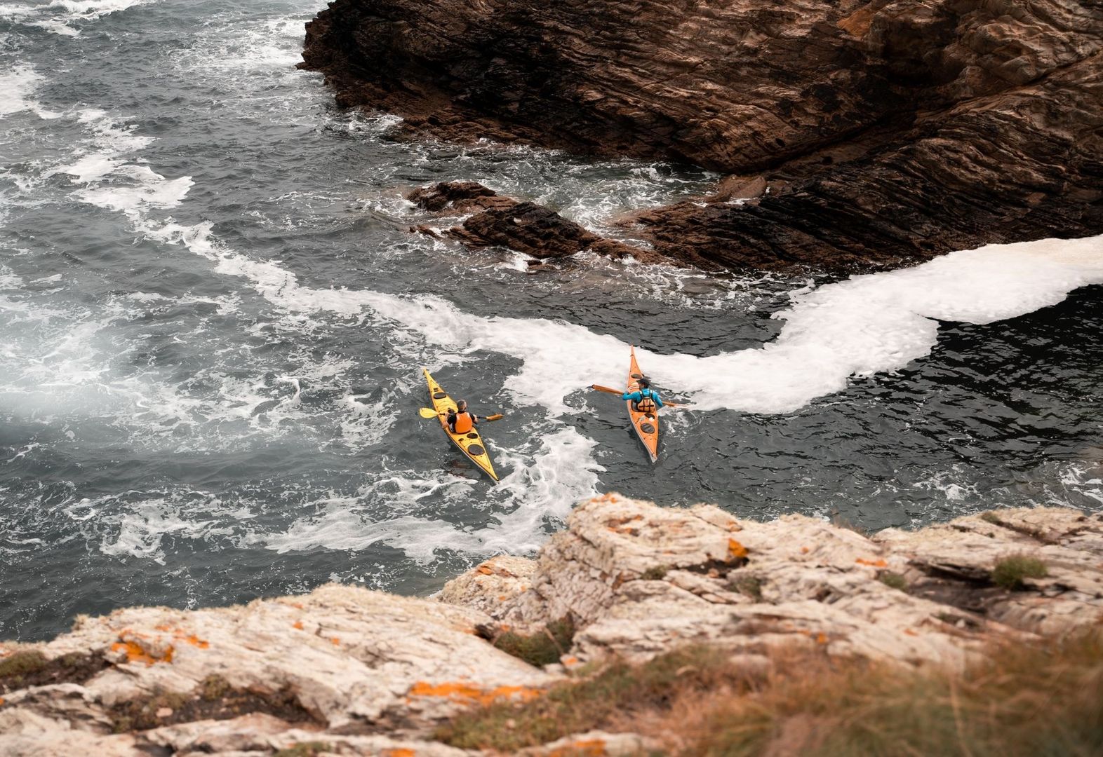 Two sea kayakers paddle through foamy water beside rocky coastal cliffs